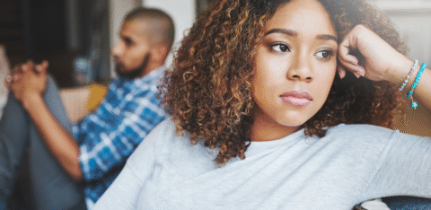 Woman sitting on a couch with a serious expression while a man sits blurred in the background, facing away.