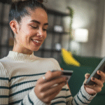 Smiling woman holding a credit card in one hand and a smartphone in the other while sitting indoors.