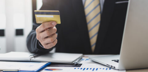 Business professional holding a credit card while sitting at a desk with a laptop and financial documents.