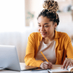 Woman using a laptop and writing in a notebook at a table.