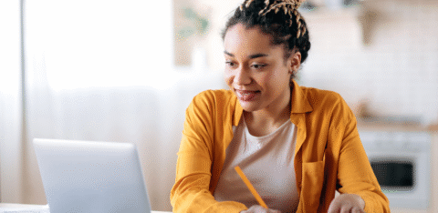 Woman using a laptop and writing in a notebook at a table.