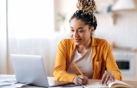 Woman using a laptop and writing in a notebook at a table.