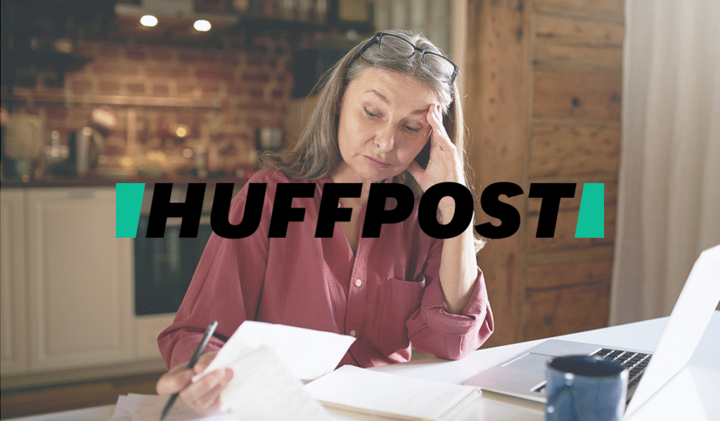 Older adult woman reviewing financial documents at home, appearing worried as she looks over papers next to an open laptop, with a HuffPost logo over the image.