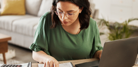 Woman reviewing paperwork at a table with a laptop and calculator.