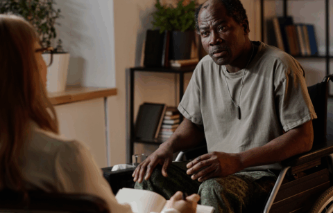Man in a wheelchair speaking with another person during a seated conversation in an office setting.