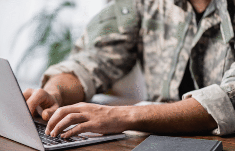 Person in military-style clothing typing on a laptop at a wooden desk.