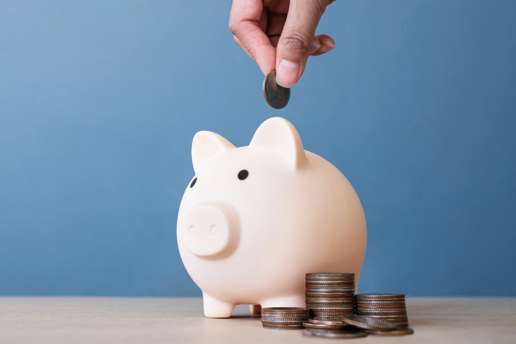Hand placing a coin into a white piggy bank, with stacks of coins beside it on a table.