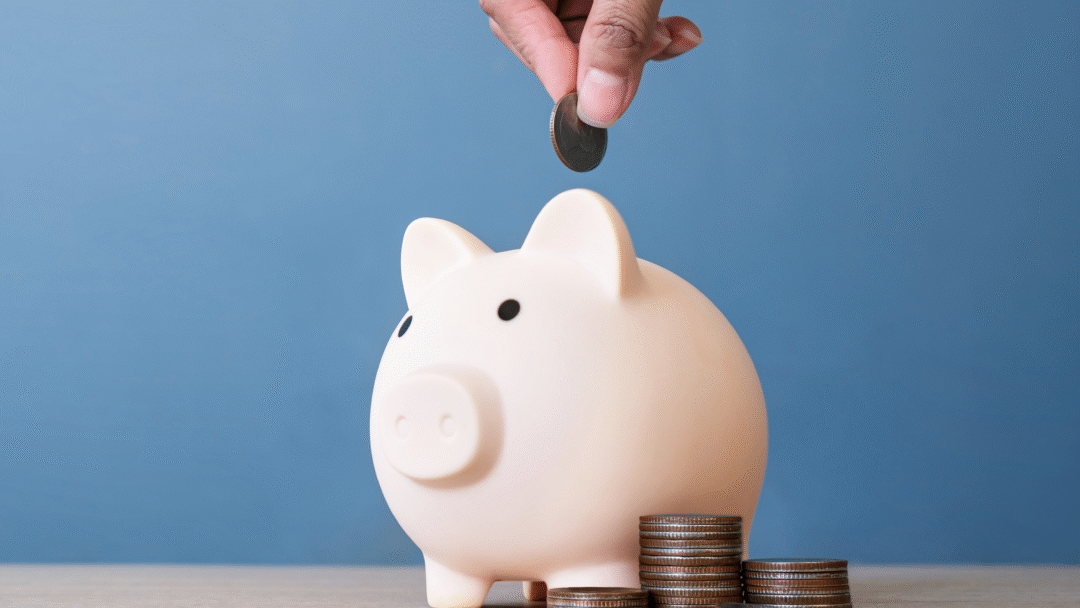 Hand placing a coin into a white piggy bank, with stacks of coins beside it on a table.