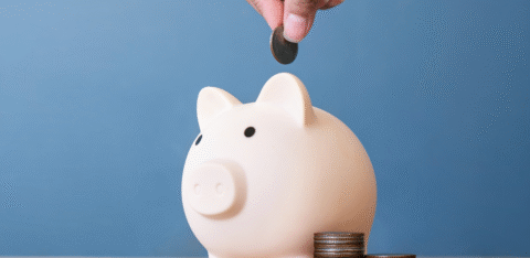 Hand placing a coin into a white piggy bank, with stacks of coins beside it on a table.
