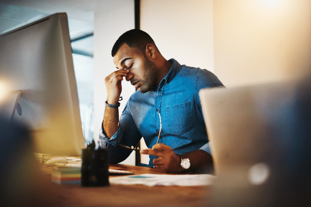 Man sitting at a desk rubbing his eyes while looking at a computer screen.