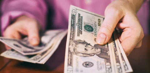 Close-up of a person holding and counting twenty-dollar bills at a table.