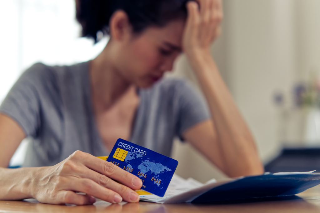 Person holding a credit card while reviewing bills at a table, with their hand on their forehead in a worried pose.
