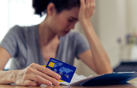 Person holding a credit card while reviewing bills at a table, with their hand on their forehead in a worried pose.