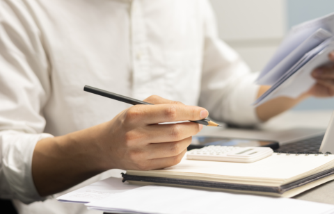 Close-up of a person holding a pencil and reviewing paperwork beside a calculator, notebook, and laptop on a desk.