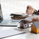 Person using a calculator near model houses and a laptop, suggesting real estate or mortgage planning