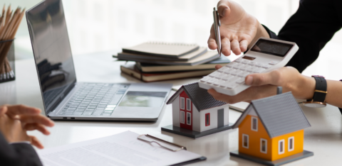 Person using a calculator near model houses and a laptop, suggesting real estate or mortgage planning