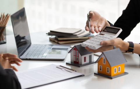 Person using a calculator near model houses and a laptop, suggesting real estate or mortgage planning