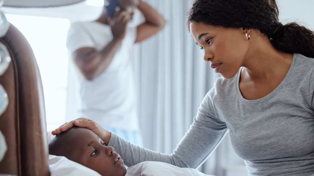 Mother checking on a sick child in bed while a man talks on the phone in the background.