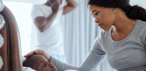 Mother checking on a sick child in bed while a man talks on the phone in the background.