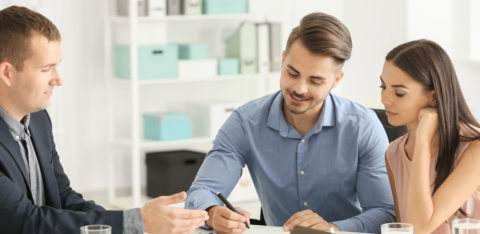 Couple signing paperwork with a professional at a desk, with a small house model in front