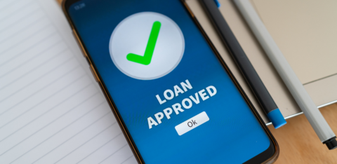 Smartphone screen displaying a green check mark and the words “Loan Approved” next to a notebook and pen.