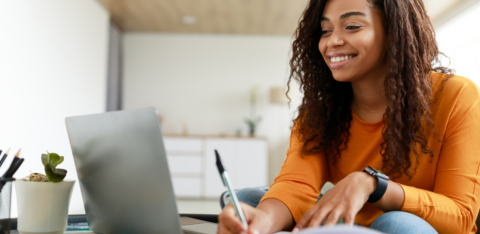 Woman smiling while using a laptop and writing in a notebook at home.