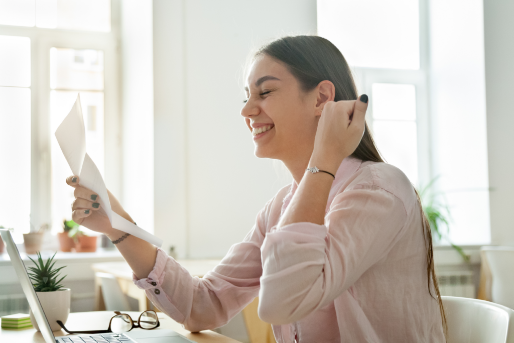 Smiling woman sitting at a desk, holding and reading a letter while looking pleased, with a laptop open in front of her.