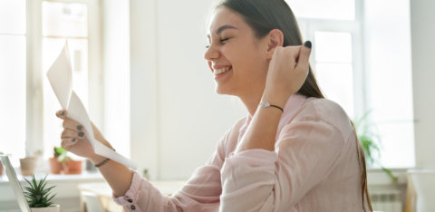 Smiling woman sitting at a desk, holding and reading a letter while looking pleased, with a laptop open in front of her.