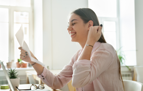 Smiling woman sitting at a desk, holding and reading a letter while looking pleased, with a laptop open in front of her.