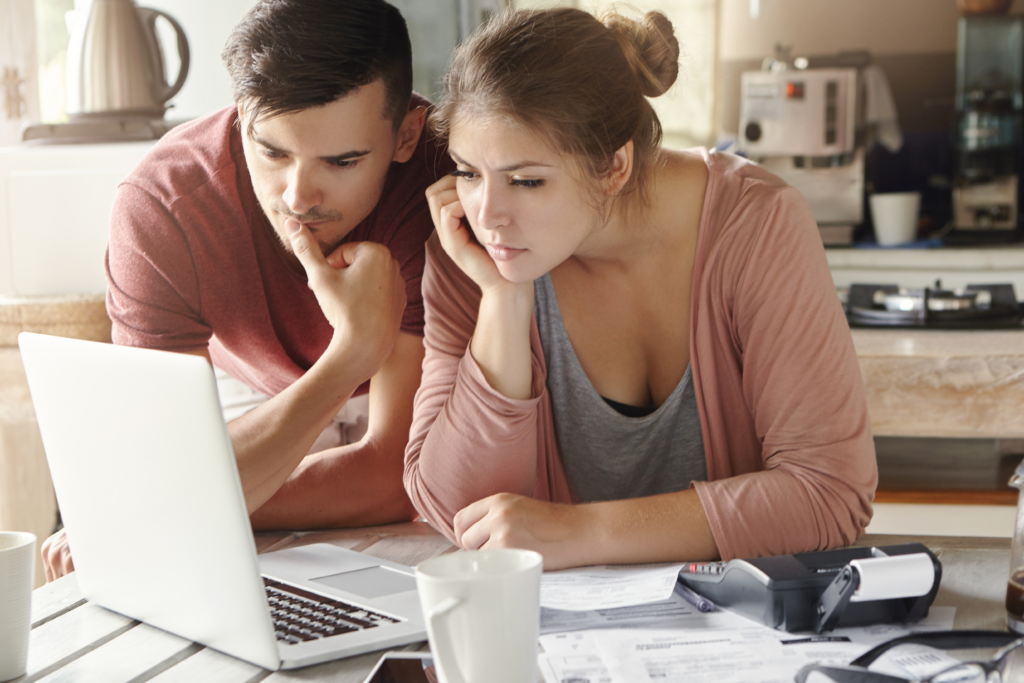 A young couple reviews bills and financial documents on a laptop at their kitchen table, with a calculator and paperwork spread out in front of them.