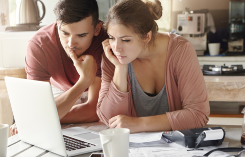 A young couple reviews bills and financial documents on a laptop at their kitchen table, with a calculator and paperwork spread out in front of them.