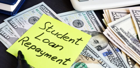 Sticky note reading “Student Loan Repayment” placed on top of U.S. dollar bills next to a calculator.