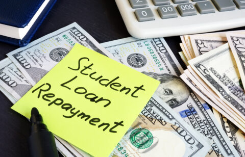Sticky note reading “Student Loan Repayment” placed on top of U.S. dollar bills next to a calculator.