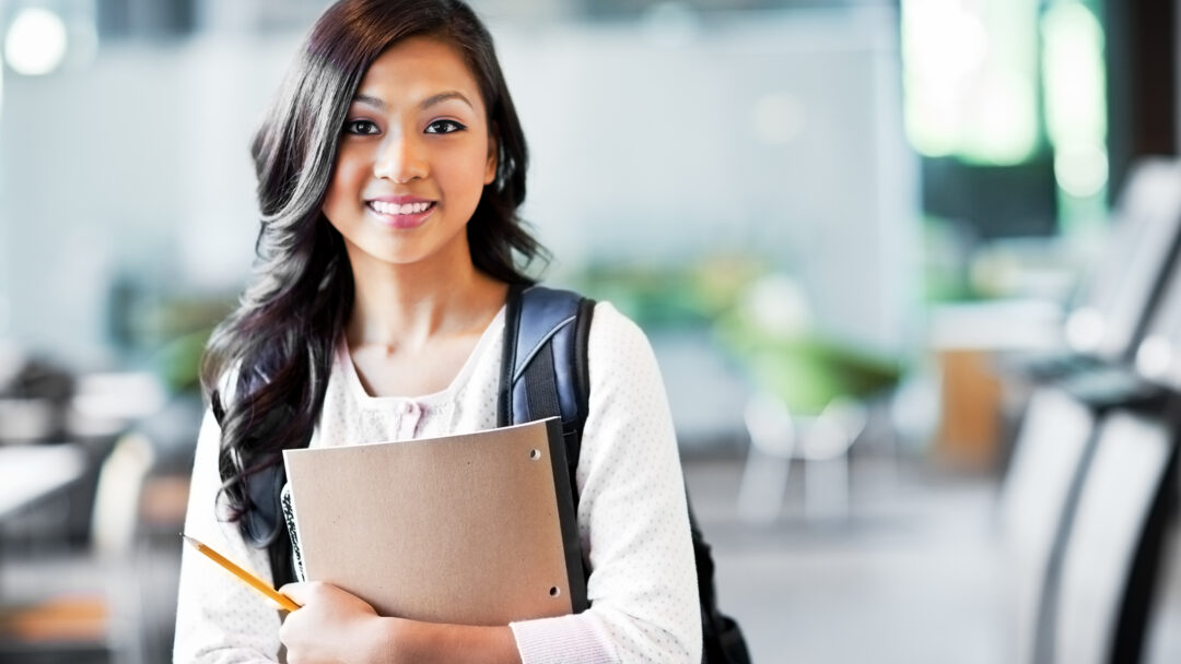 Young woman with a backpack holding a notebook and pencil, smiling in a school setting.