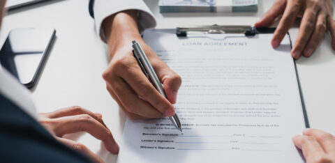 A person signs a loan agreement on a clipboard while another person points at the document, with a stack of cash nearby.