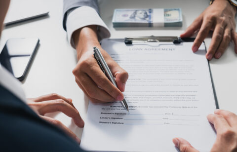 A person signs a loan agreement on a clipboard while another person points at the document, with a stack of cash nearby.