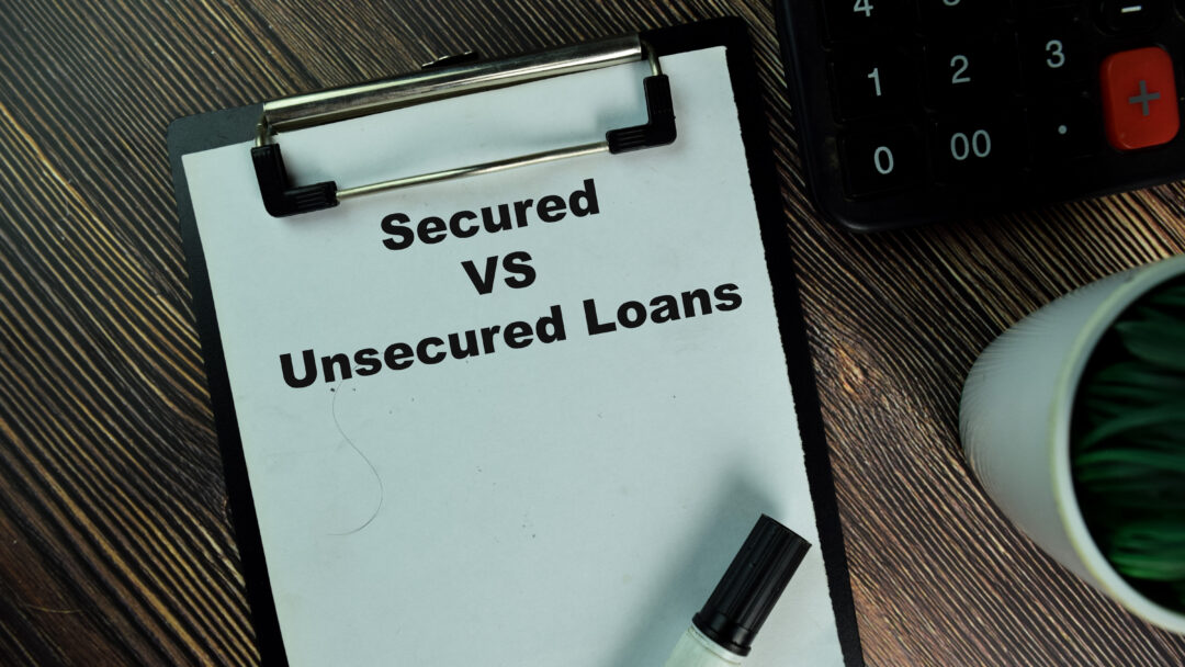 Clipboard with the words “Secured vs Unsecured Loans” next to a calculator and pen on a desk.