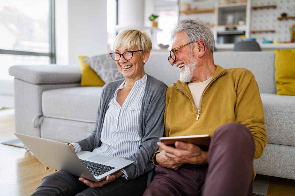 Older couple sitting on the floor at home, smiling while using a laptop and tablet.