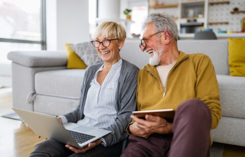Older couple sitting on the floor at home, smiling while using a laptop and tablet.