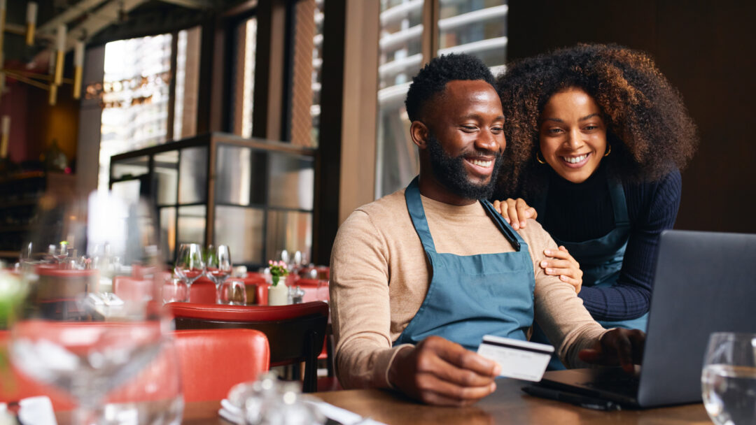 Pair of small business workers reviewing something online with a laptop and credit card inside a dining area.