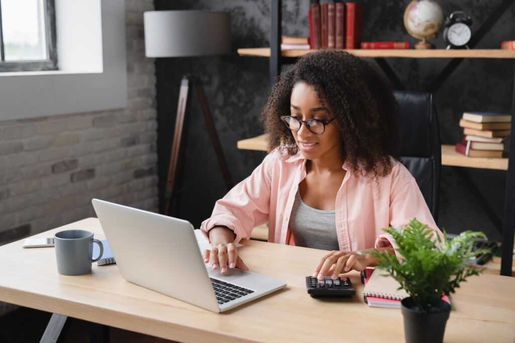 Woman working at a desk with a laptop and calculator, managing finances in a home office.