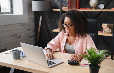 Woman working at a desk with a laptop and calculator, managing finances in a home office.