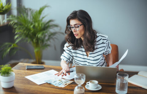 Woman reviewing paperwork and cash at a table while using a laptop.