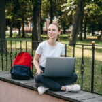 Student sitting outdoors with a laptop on a ledge, with a backpack beside her.