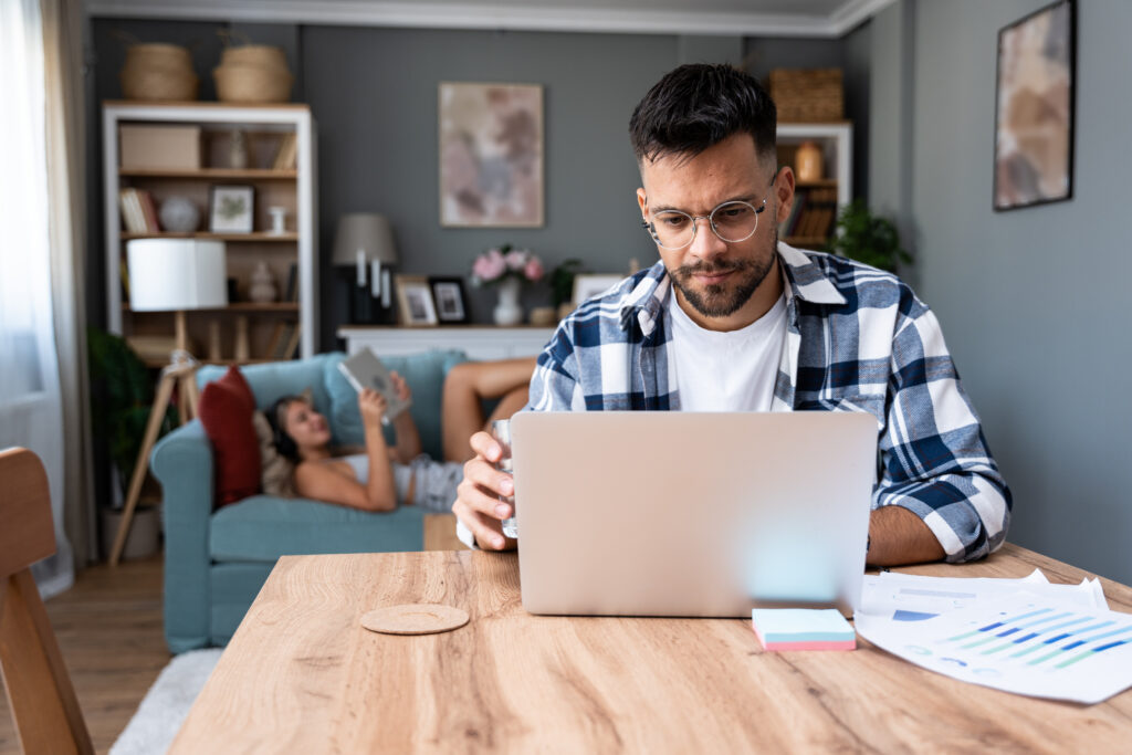 Man working on a laptop at a table while holding a glass of water, with a woman relaxing on a couch in the background.