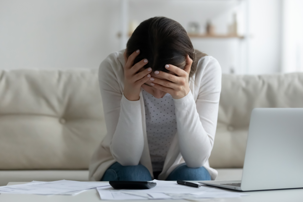 Person sitting on a couch with head in hands, surrounded by bills, a calculator, and a laptop.