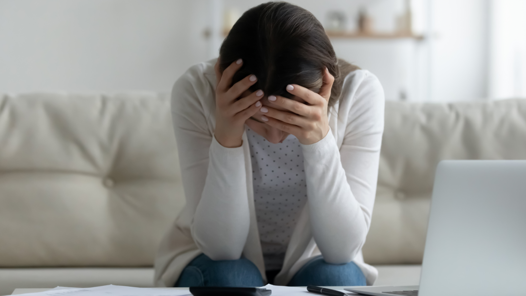 Person sitting on a couch with head in hands, surrounded by bills, a calculator, and a laptop.