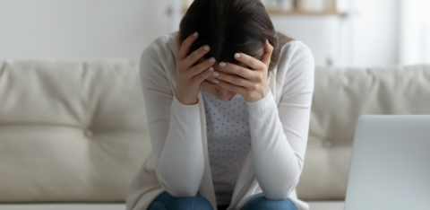 Person sitting on a couch with head in hands, surrounded by bills, a calculator, and a laptop.