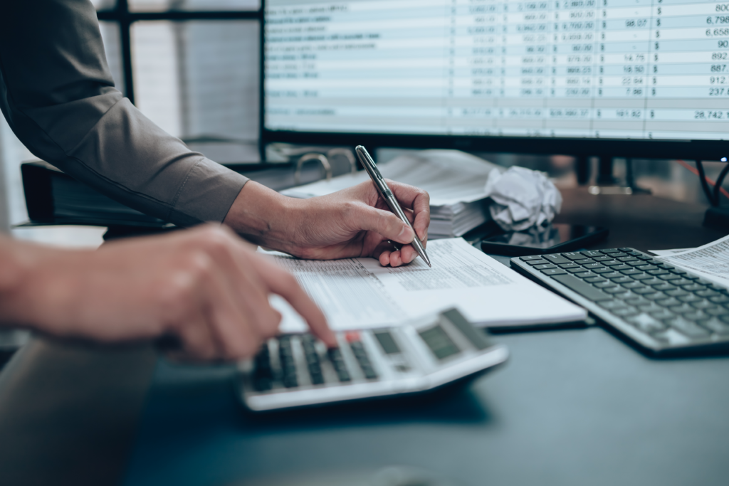 Person using a calculator and writing on paperwork at a desk with financial data on a computer screen