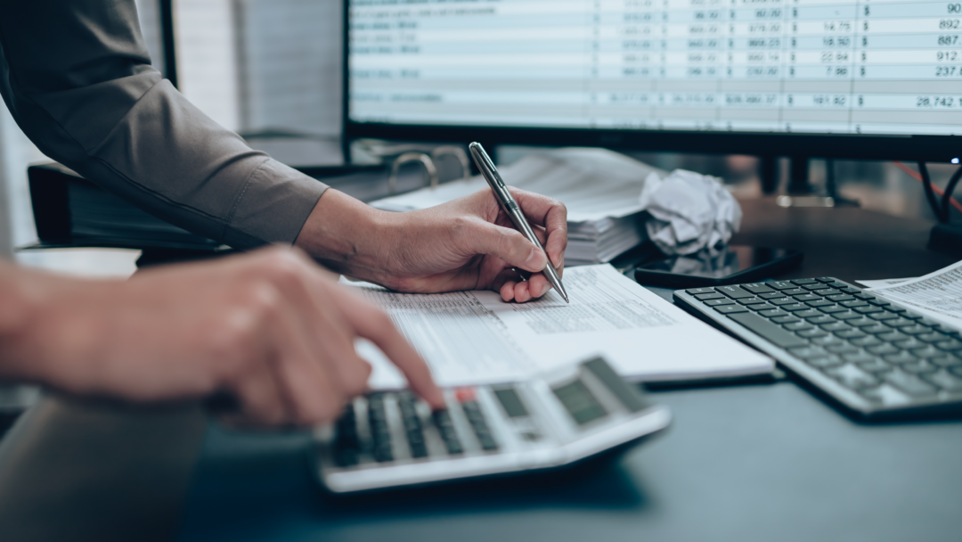 Person using a calculator and writing on paperwork at a desk with financial data on a computer screen
